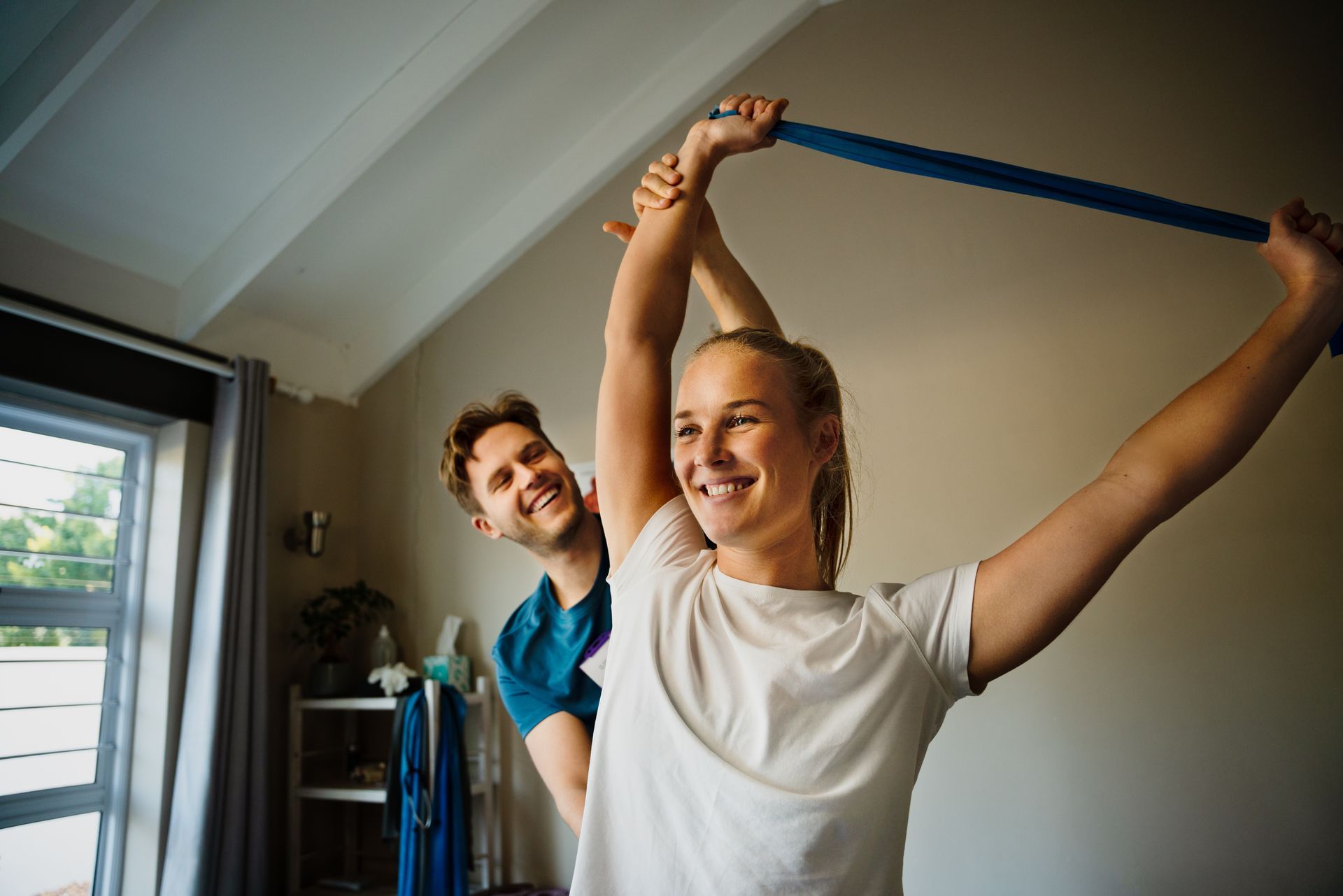 Two people exercising indoors with a blue resistance band, smiling near a window.
