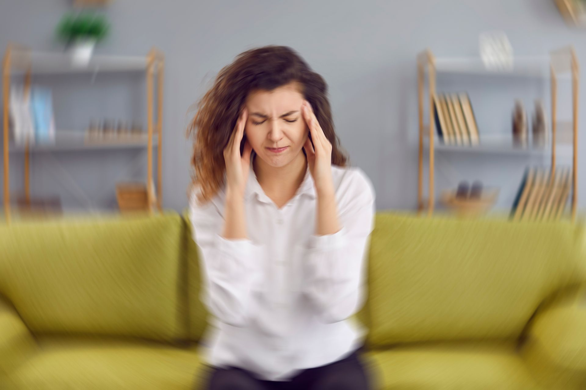 Person sitting on a green couch, holding their head in a blurred living room, looking distressed