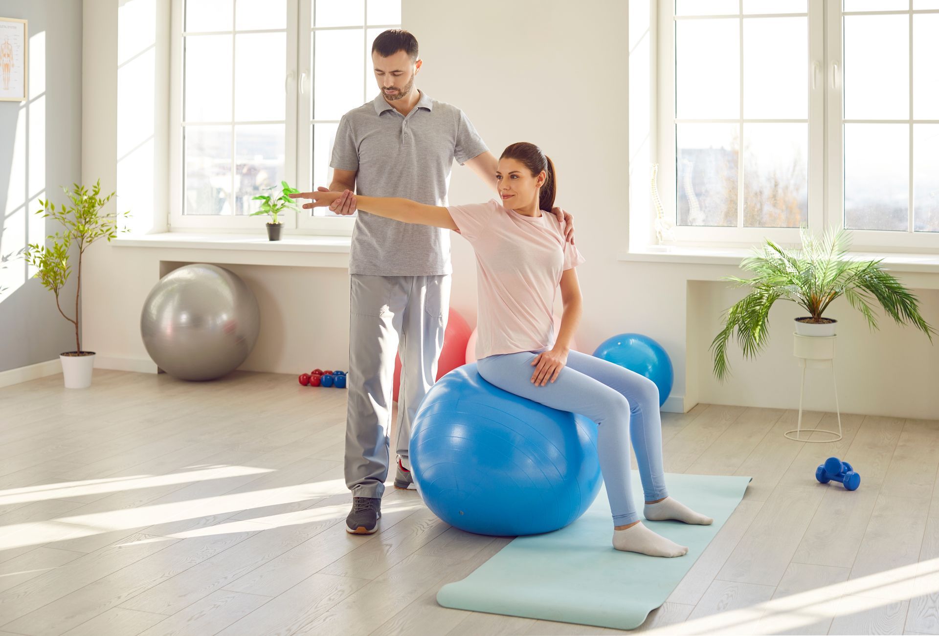 Person assisting another doing balance exercise on a blue ball in a bright studio room