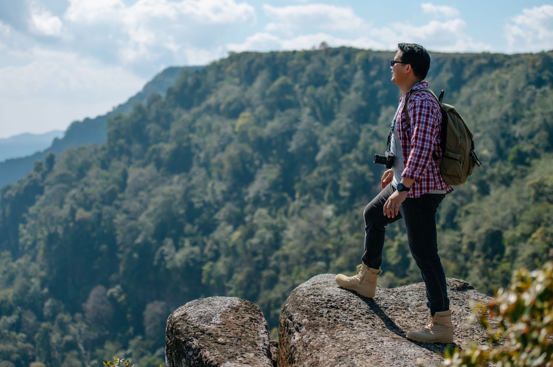 Hiker standing on rocky outcrop with backpack, overlooking forested mountains under a cloudy sky