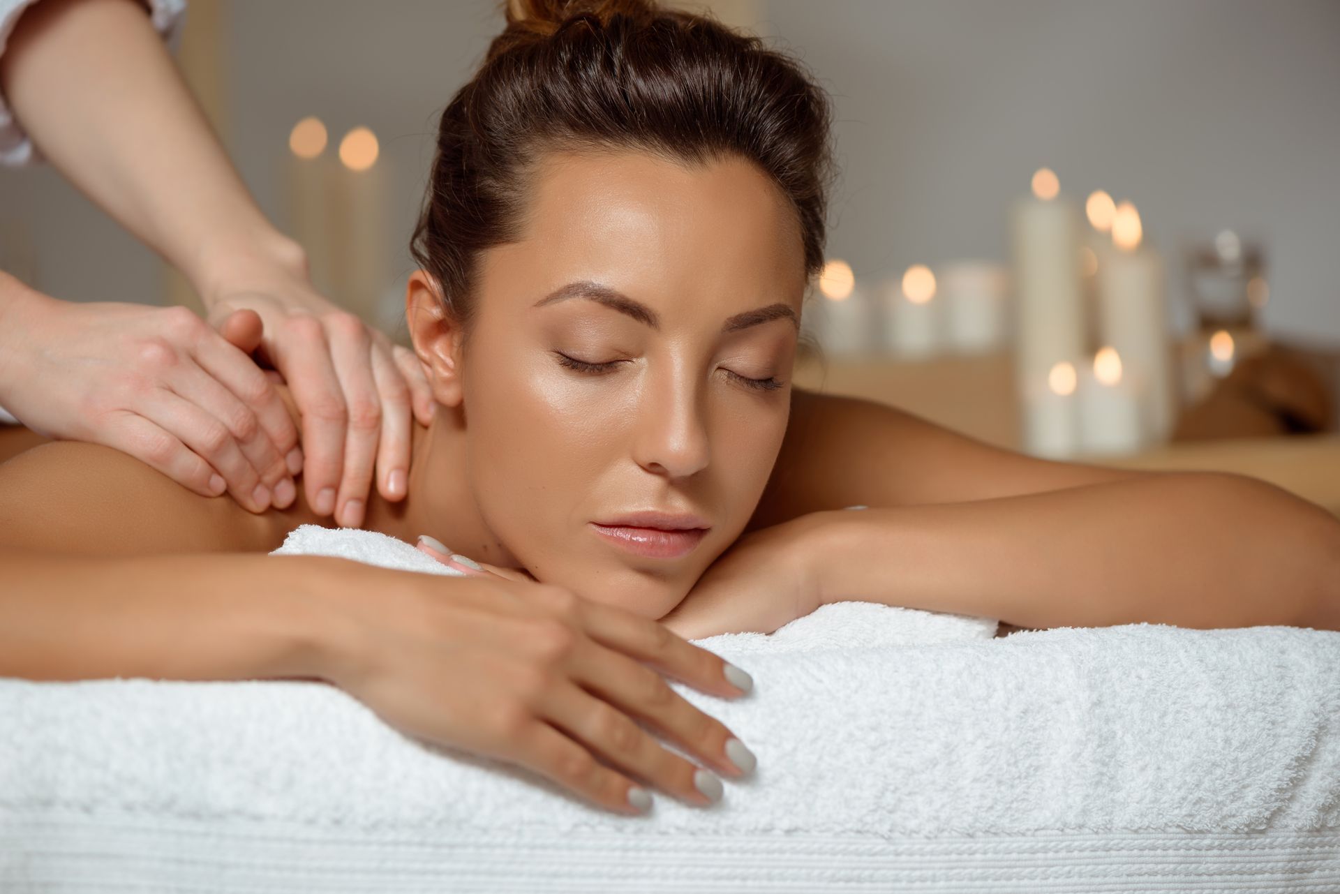 Person receiving a massage with eyes closed on a white towel in a spa, with soft candlelight in the background