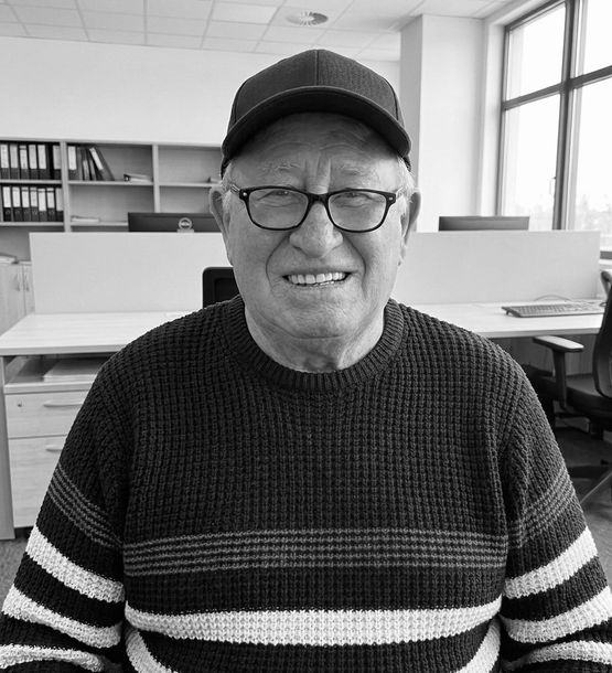 Persona sorridente con occhiali e berretto, che indossa un maglione a righe, seduta in una stanza simile a un ufficio.