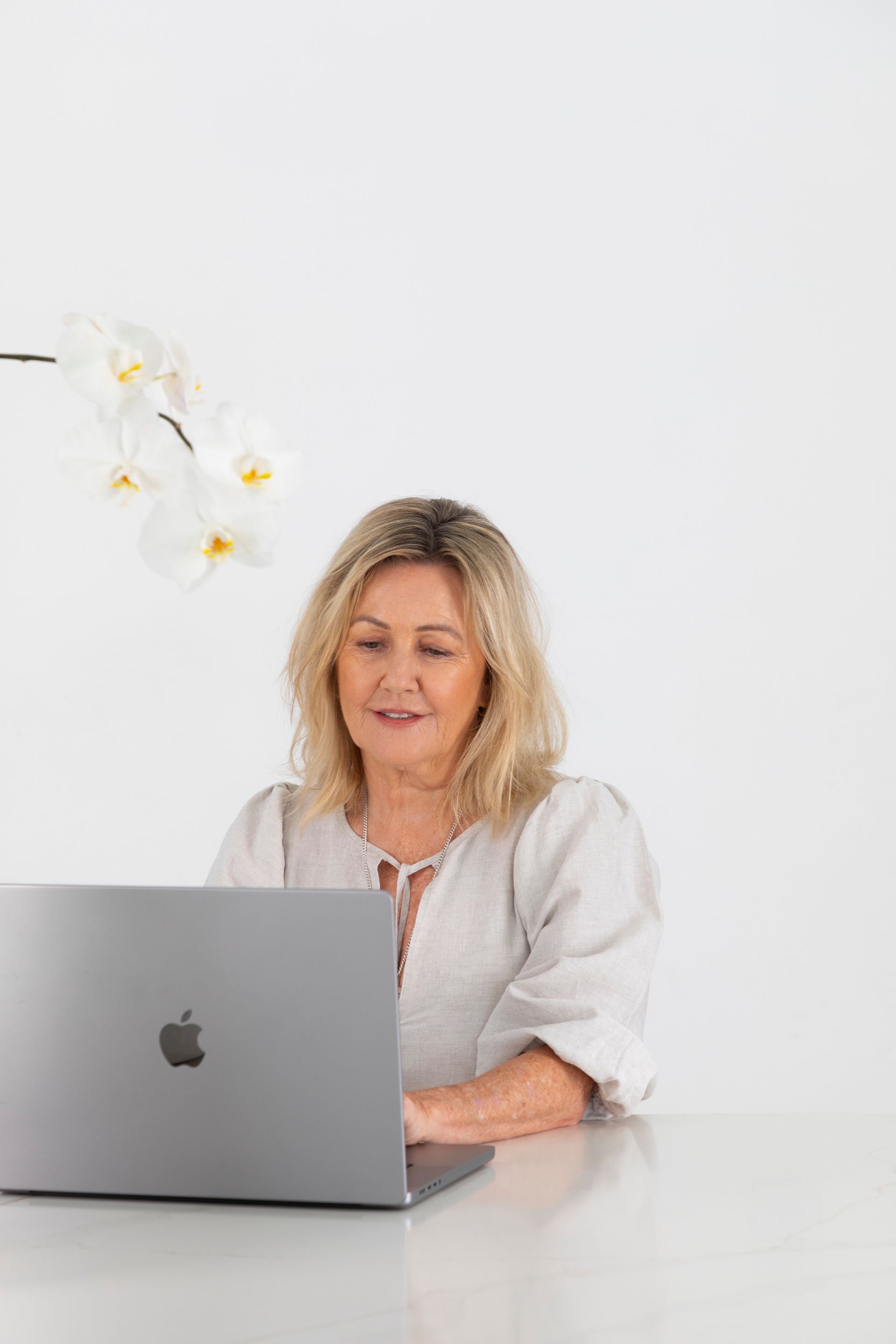 Woman with blonde hair smiles, using a laptop at a white table with orchids in background.