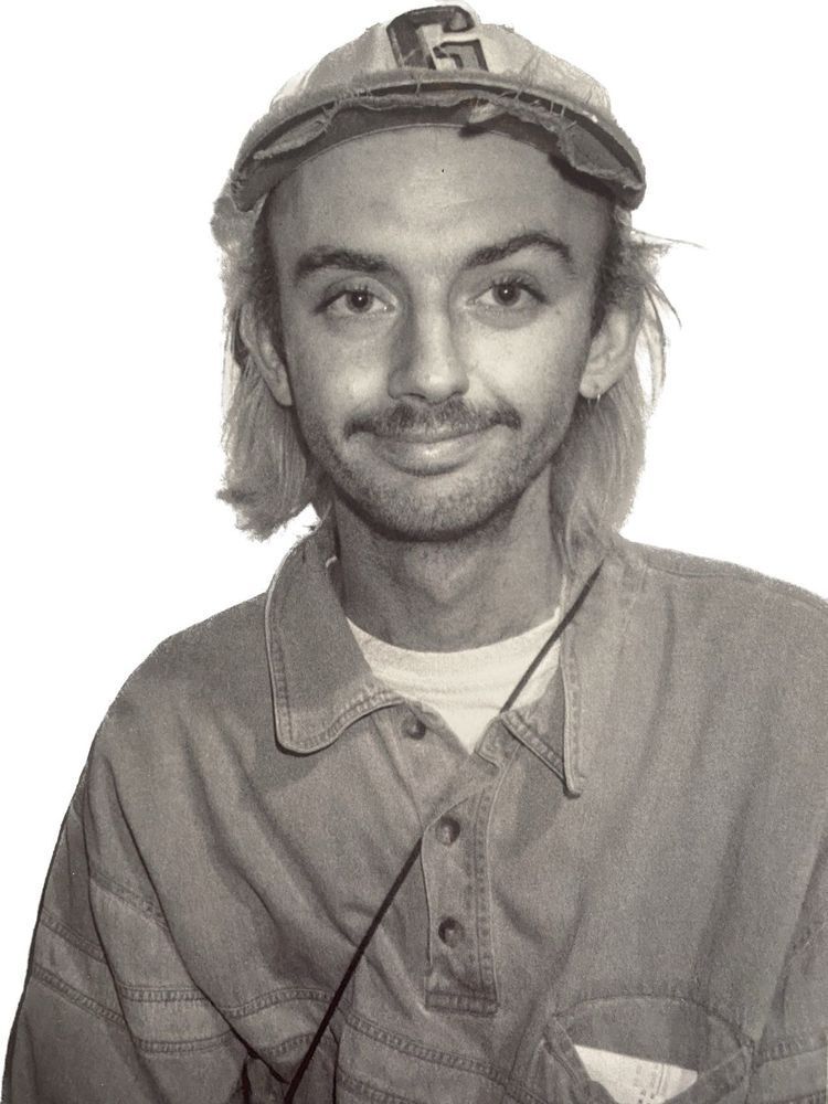 Man with long blonde hair and a cap, smiling, wearing a denim shirt.