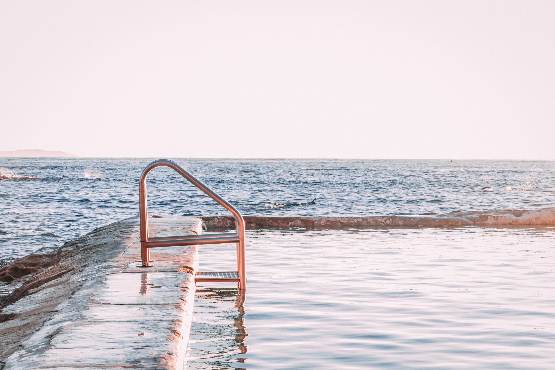 Blue ocean pool reflecting a pink sky and ocean with surfers.
