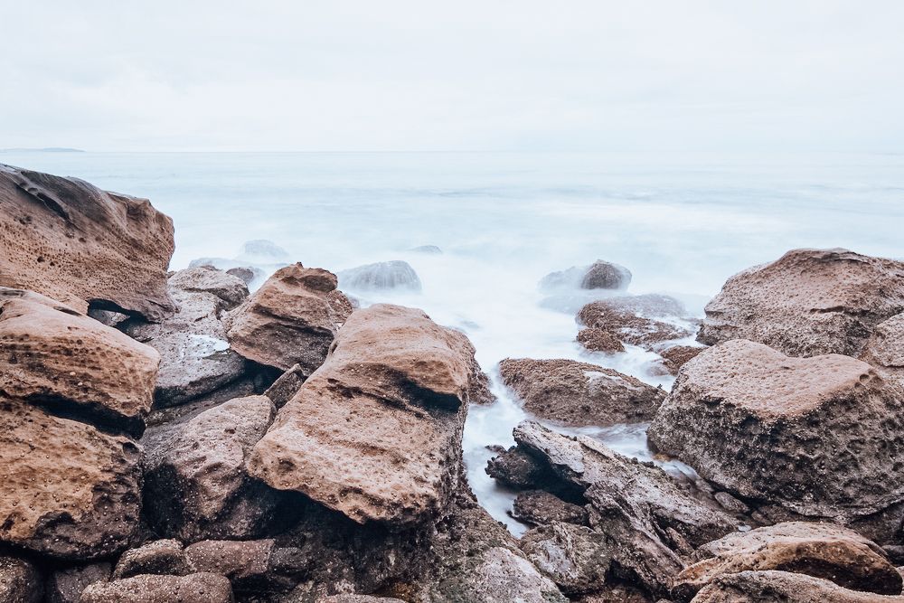 Rocky shoreline, light brown rocks with waves washing over them, overcast sky.