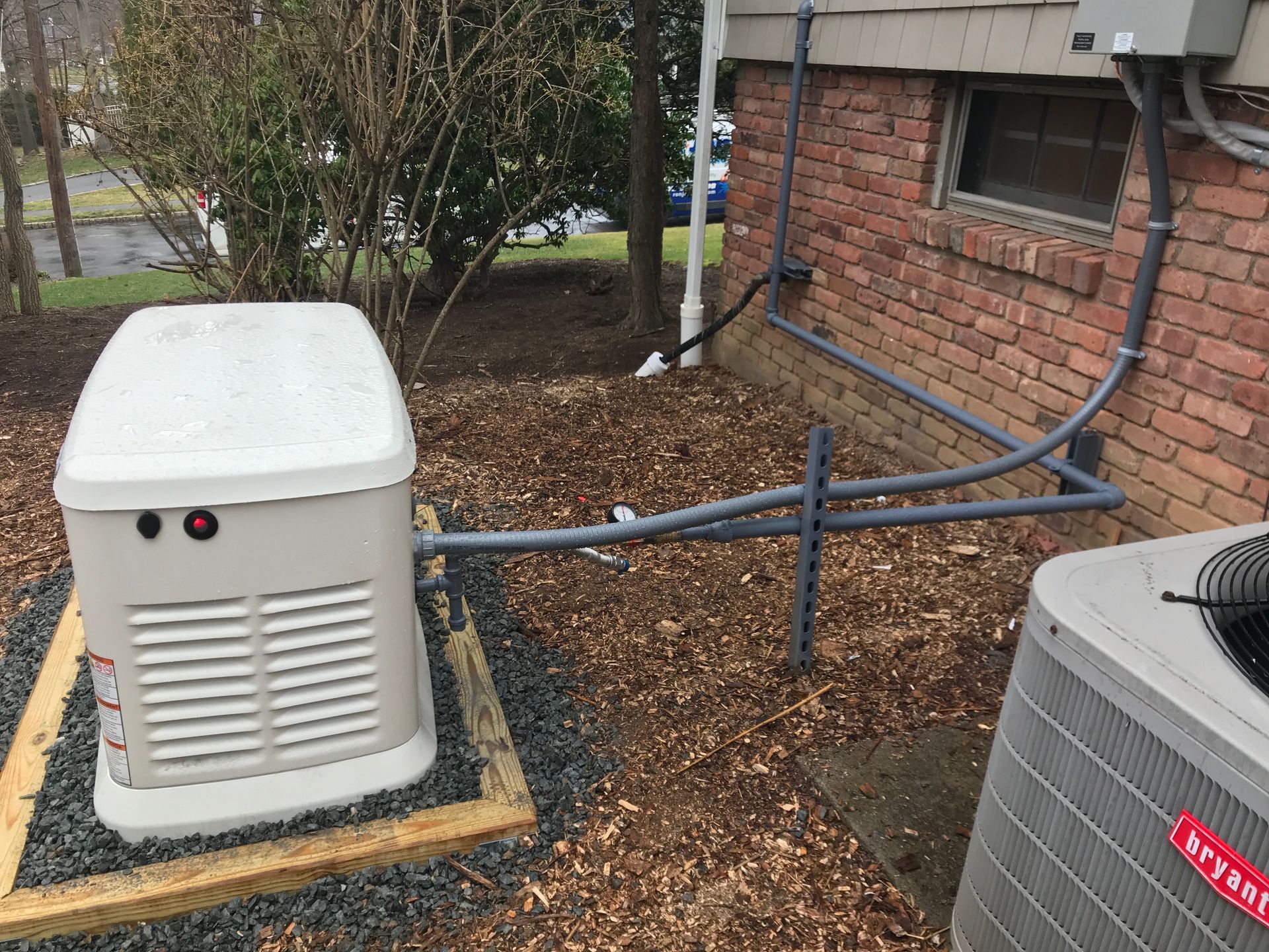 Outdoor HVAC generator beside a brick building, connected by gray conduit and surrounded by mulch.