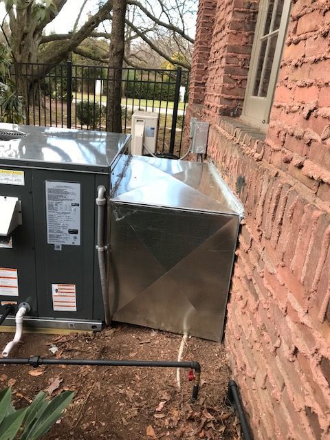 Outdoor HVAC units beside a brick house, with a metal duct enclosure in a yard.