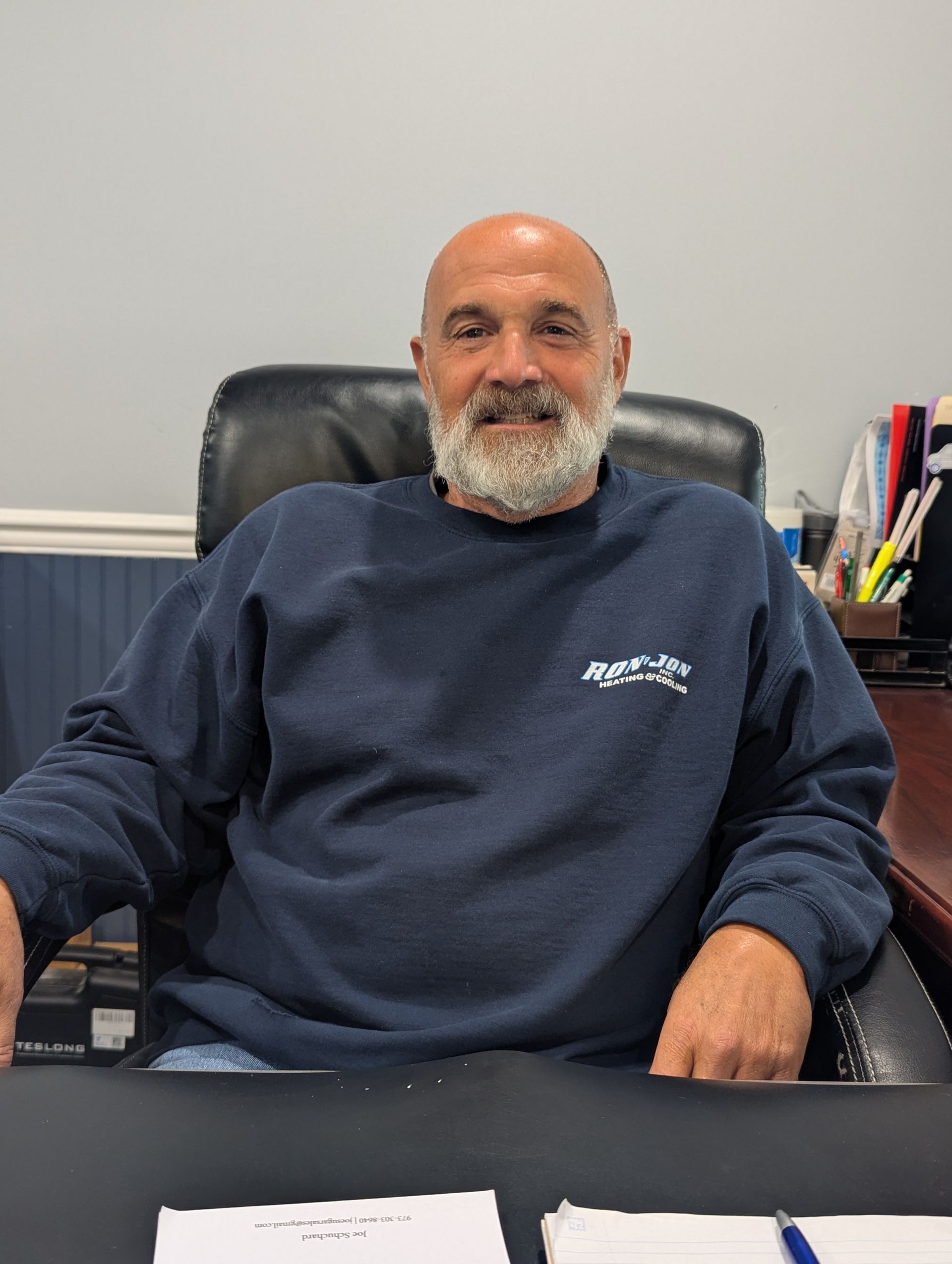 Man sitting in an office chair at a desk, wearing a navy sweatshirt, smiling slightly.