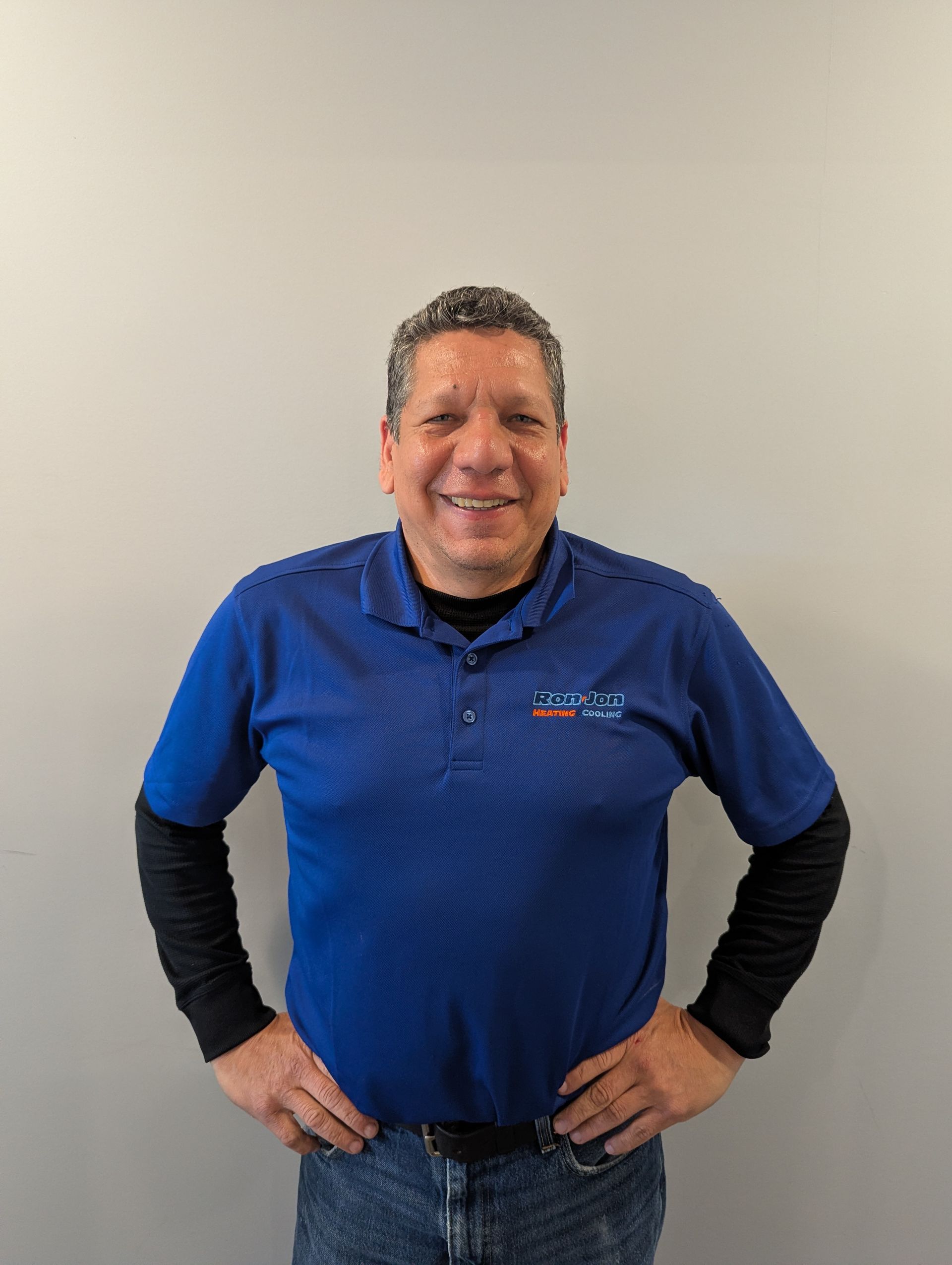 Person smiling in a blue polo shirt and black sleeves, standing with hands on hips against a plain wall