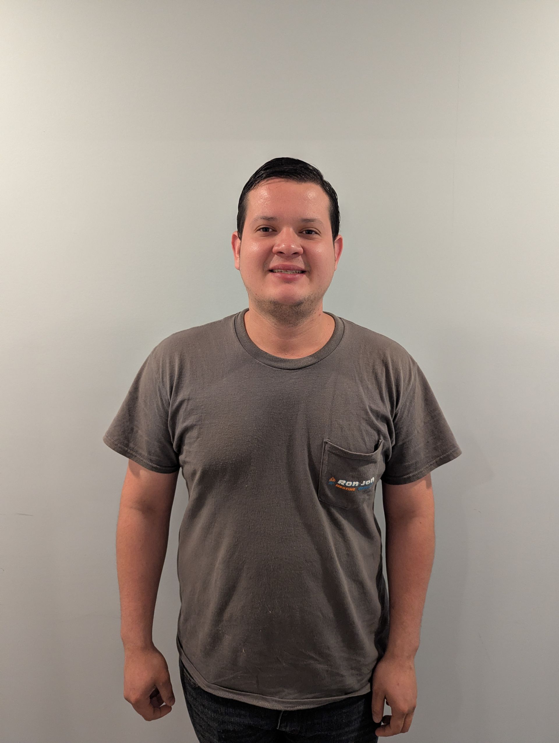 A man in a gray T-shirt stands against a plain white wall, smiling at the camera.