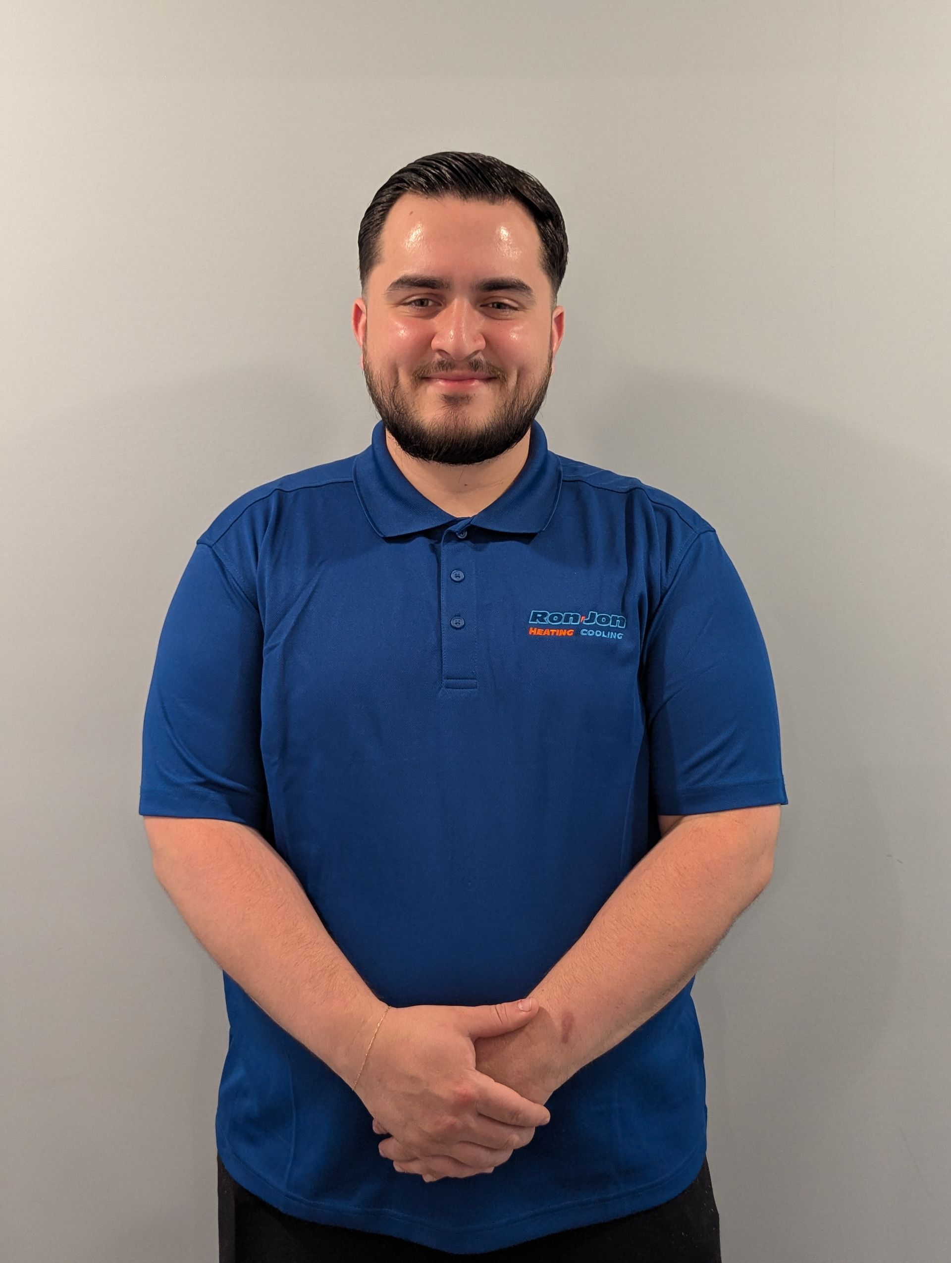 Man standing against a plain wall, wearing a blue polo shirt and clasping his hands in front of him.