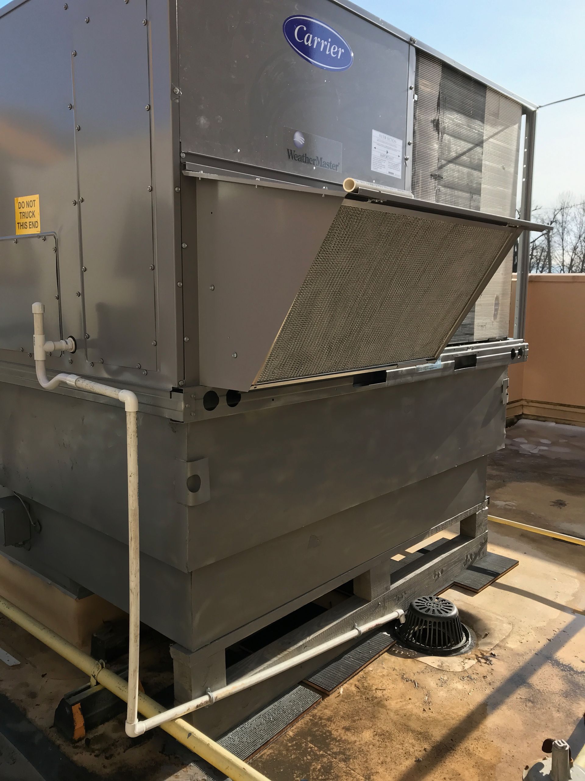 Large gray HVAC unit outdoors with a bent metal panel and pipe connections on a concrete pad.