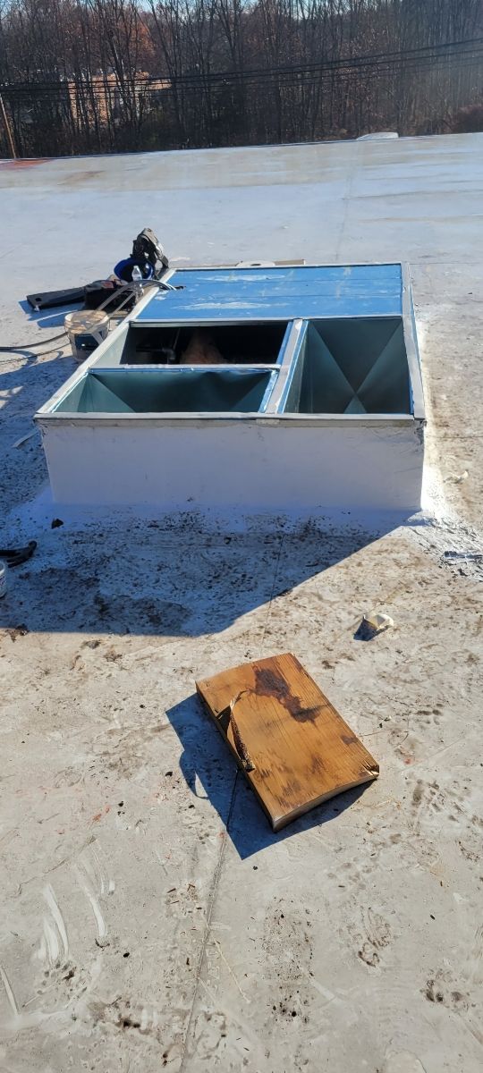 White rooftop vent box with open blue lid on snow-covered roof, with a small brown board in the foreground