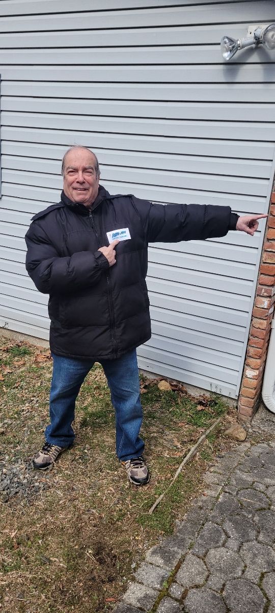 Man in a black jacket points beside a gray-sided building outdoors.