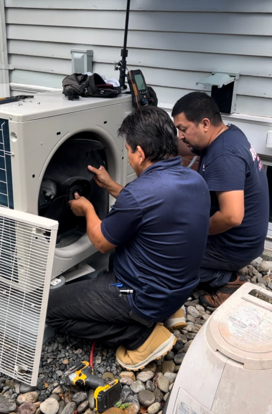 Two technicians kneel beside an outdoor HVAC unit, inspecting its open fan compartment.
