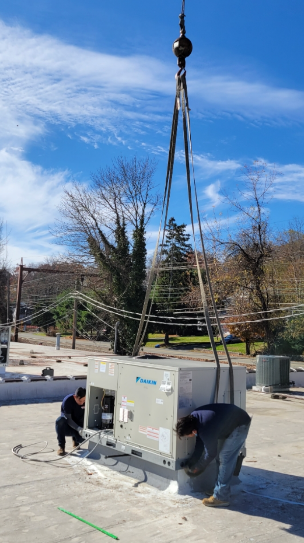 Crane lifts a large beige HVAC unit while two workers guide it outdoors.