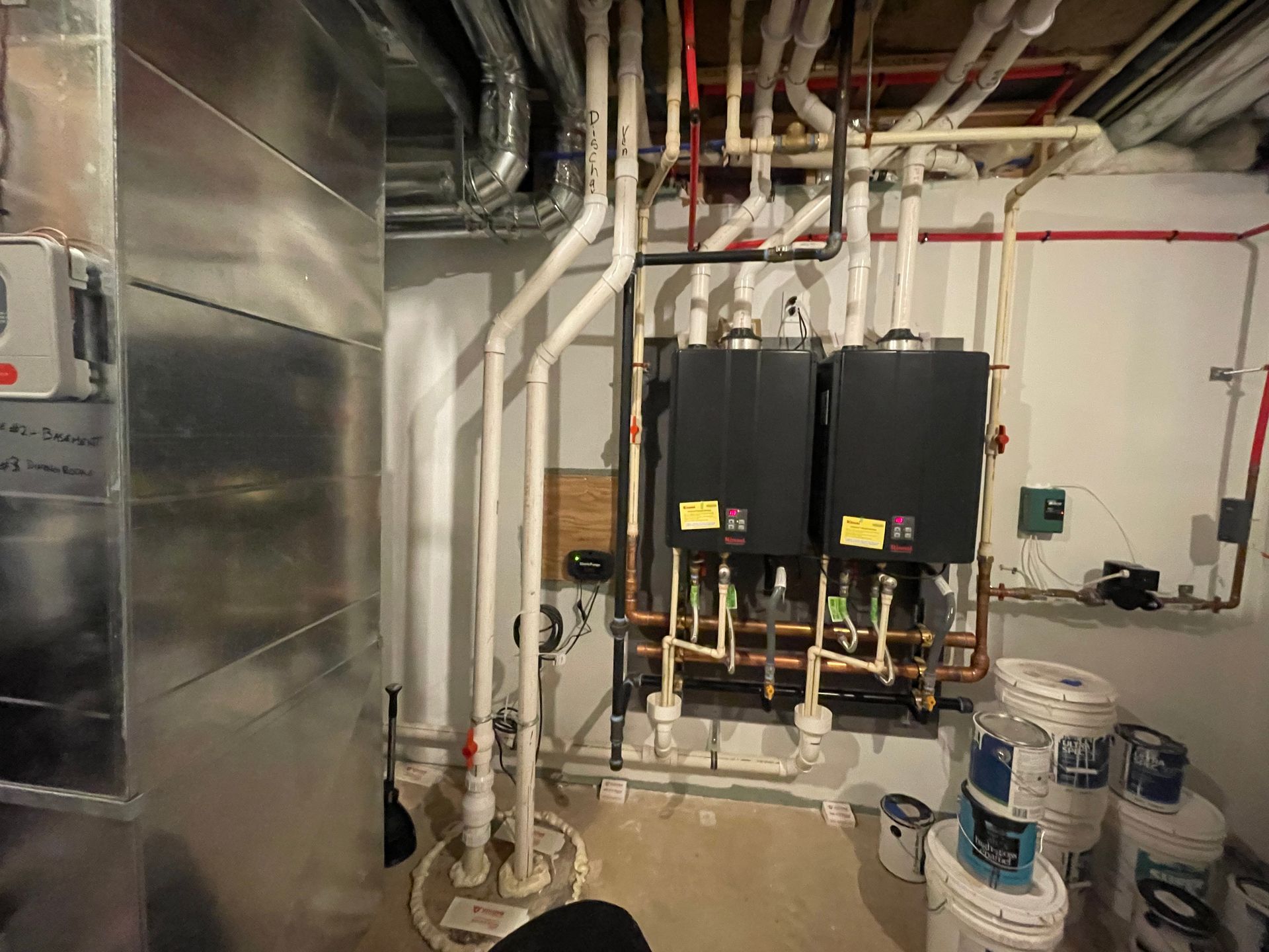 Industrial utility room with pipes, ductwork, two black tank units, and stacked buckets on the floor