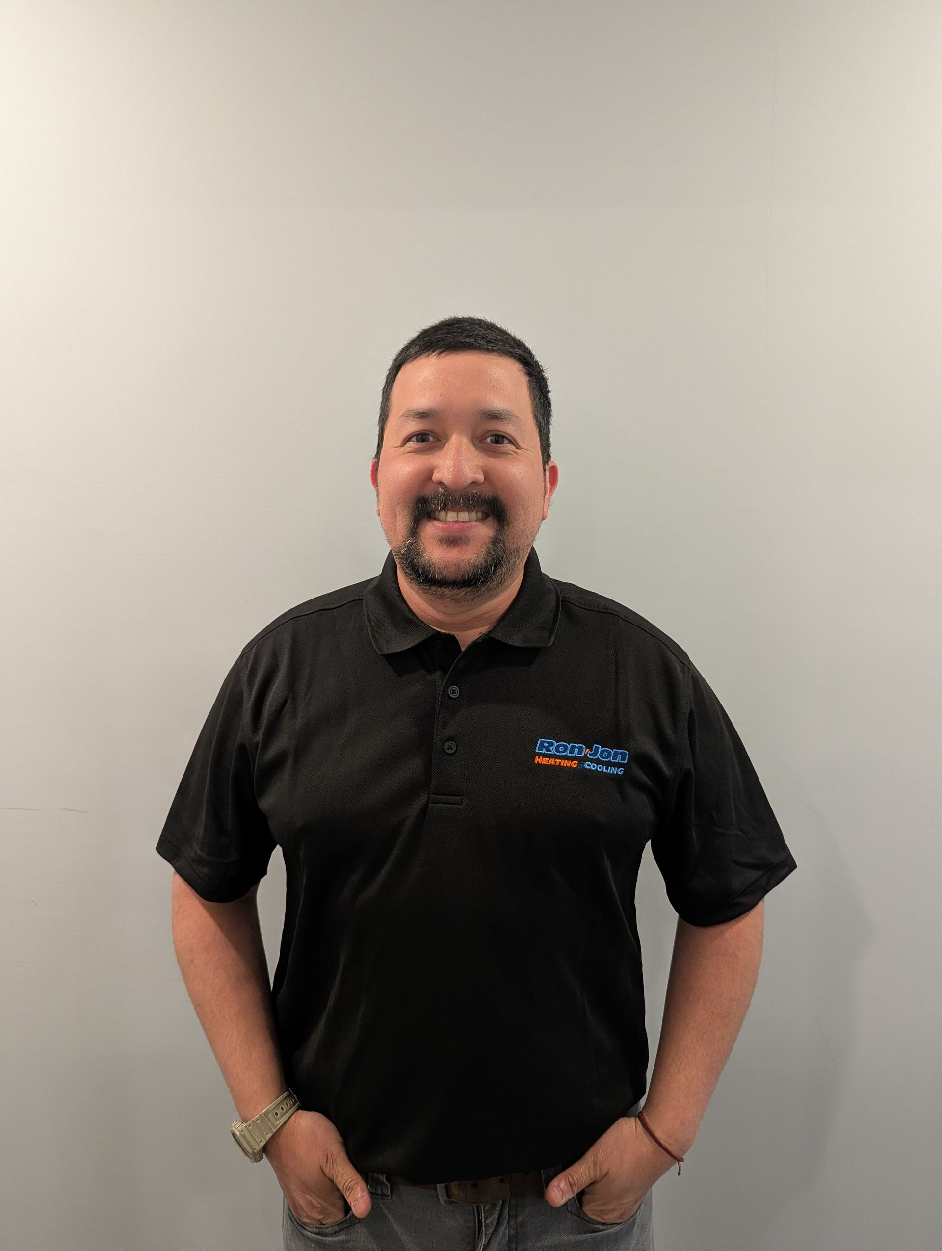Man standing against a plain gray wall, smiling, wearing a black polo shirt and jeans, hands in pockets