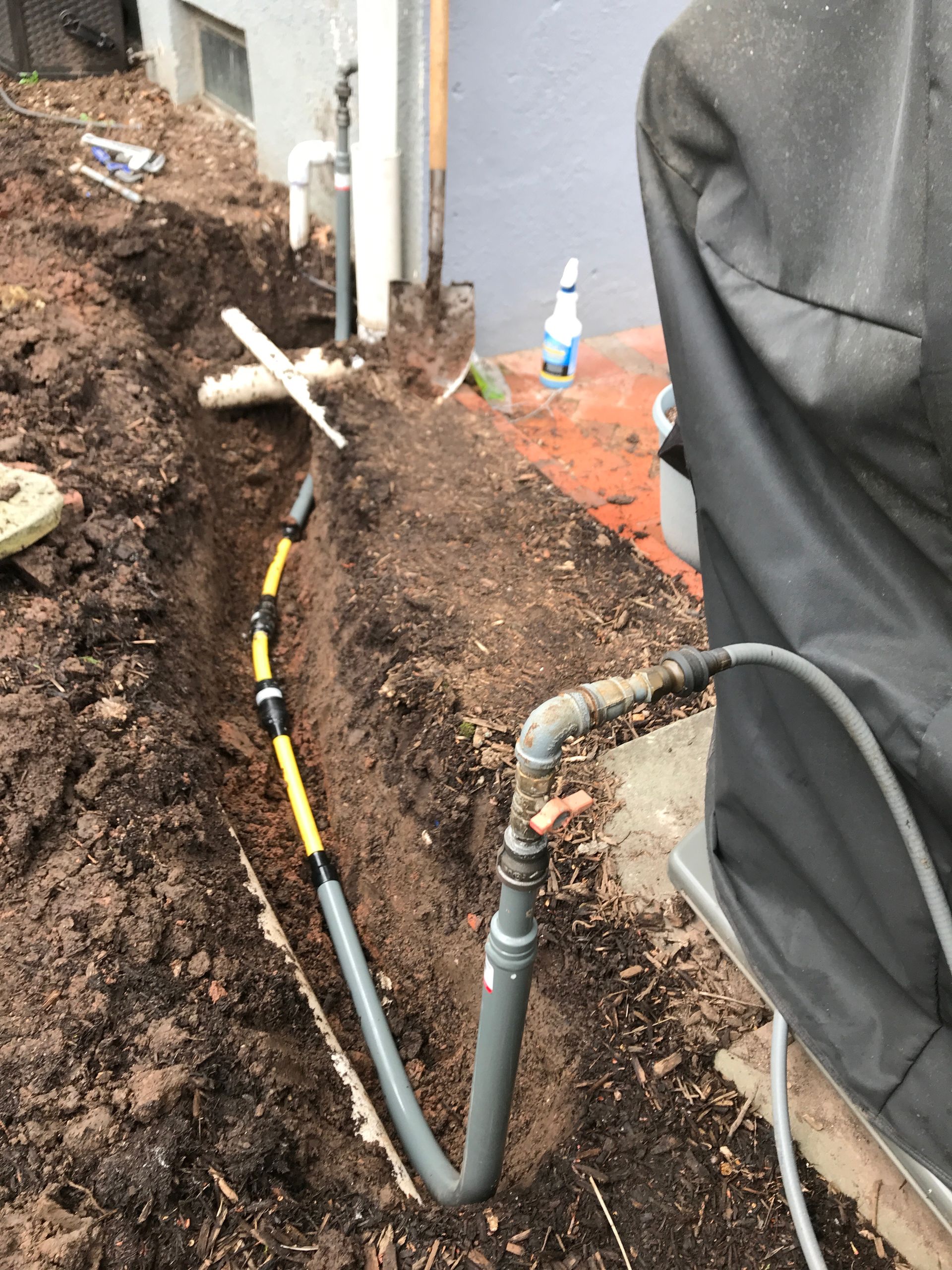 Person working near exposed utility pipes in a muddy trench beside a building