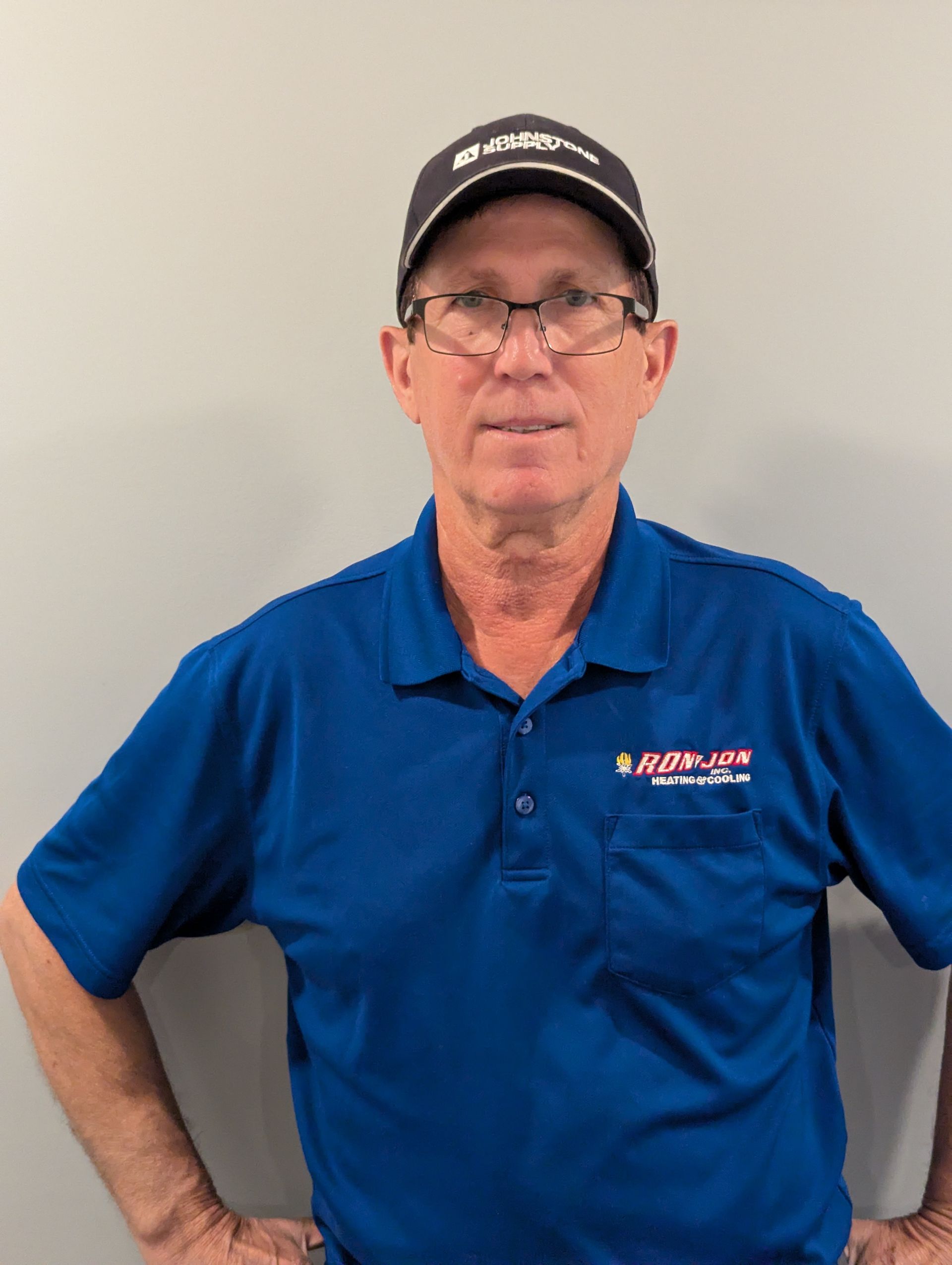 Man in a blue polo and black cap standing against a plain wall with one hand on his hip