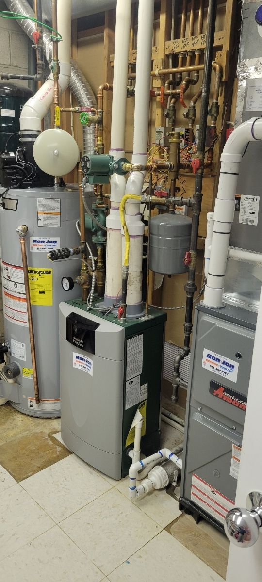 Basement utility room with furnaces, water heaters, pipes, and gas lines against a brick wall.