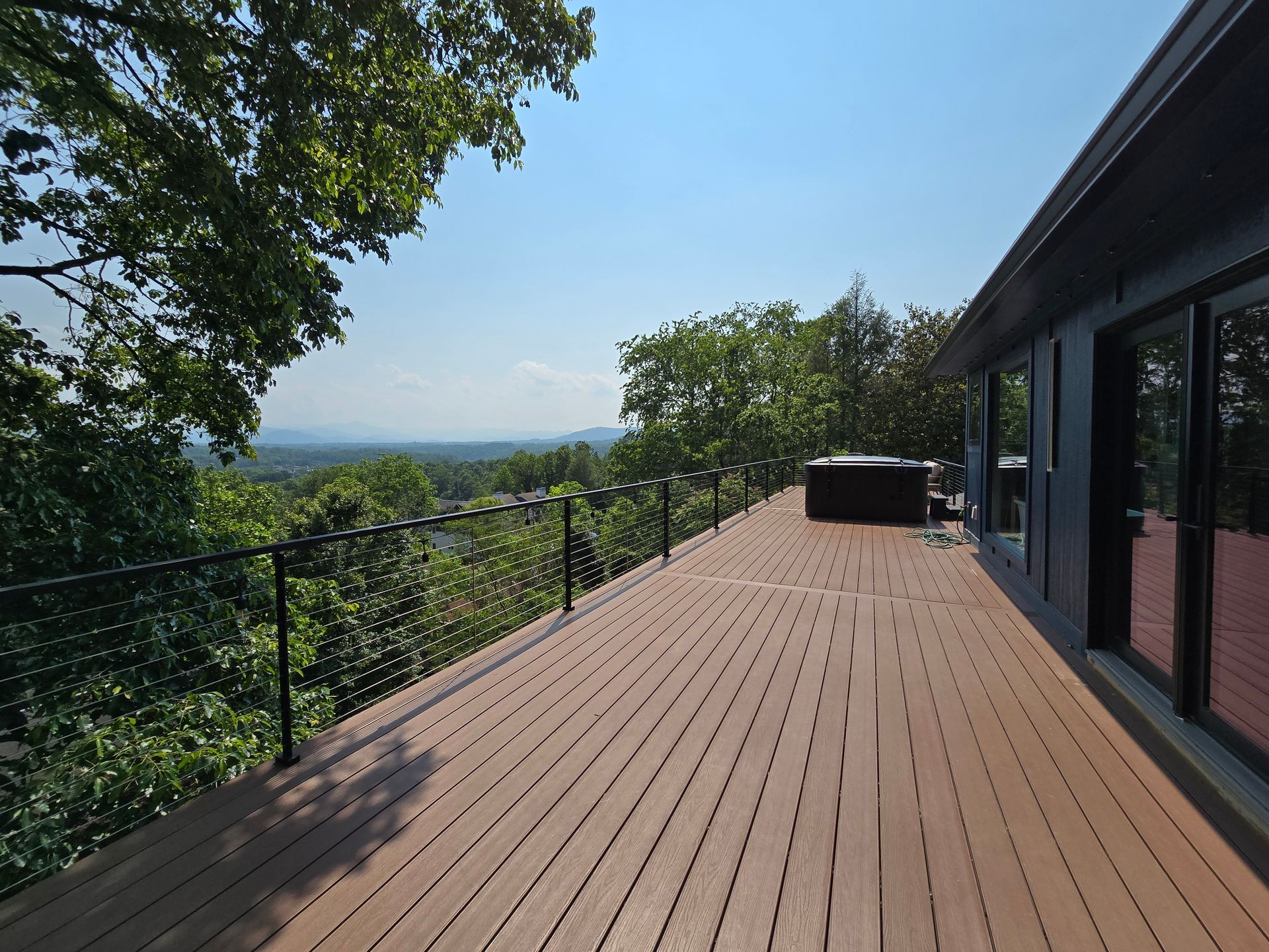 Wooden deck with black railing overlooking a distant mountain range; hot tub and modern house.
