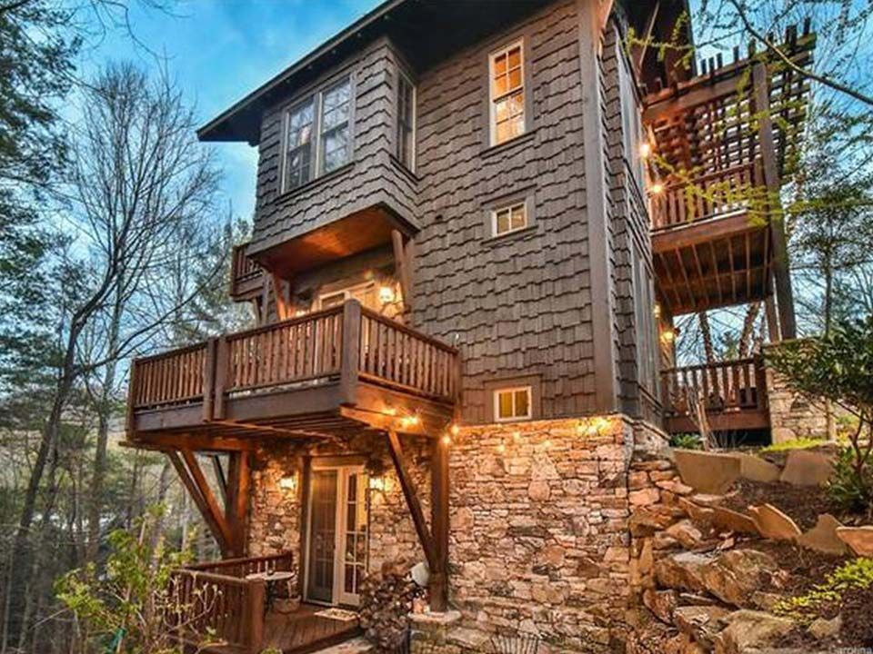 Rustic multi-story cabin nestled in a wooded area; brown shingles, stone foundation, wooden balconies, string lights.