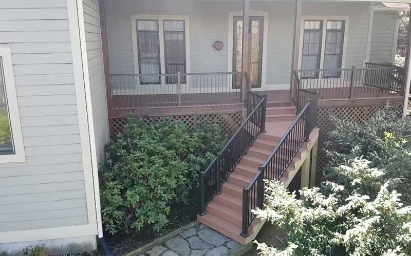 Exterior of a house with a porch and stairs leading to the front door. Wooden deck, black railing, and shrubbery.