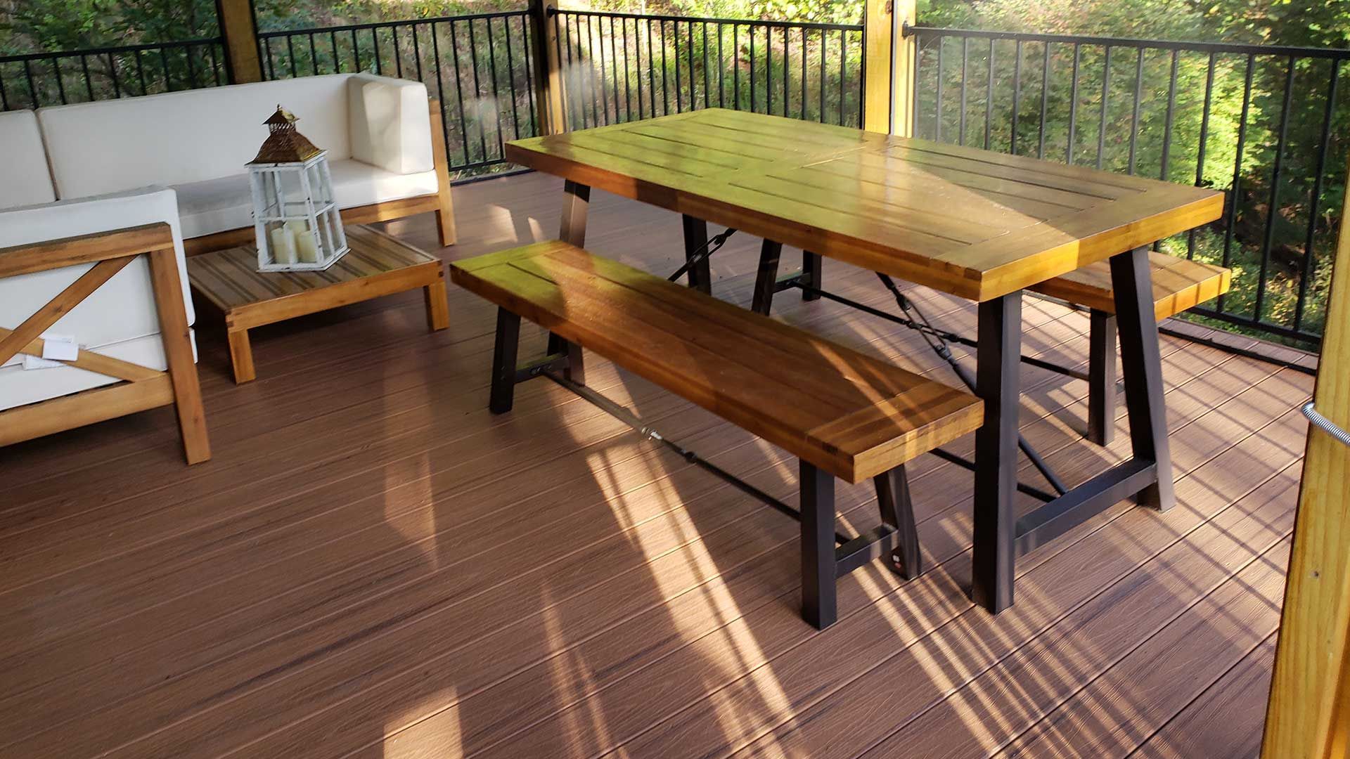 Outdoor deck with wooden furniture: sofa, table, and benches. Brown and black tones with sunlight.