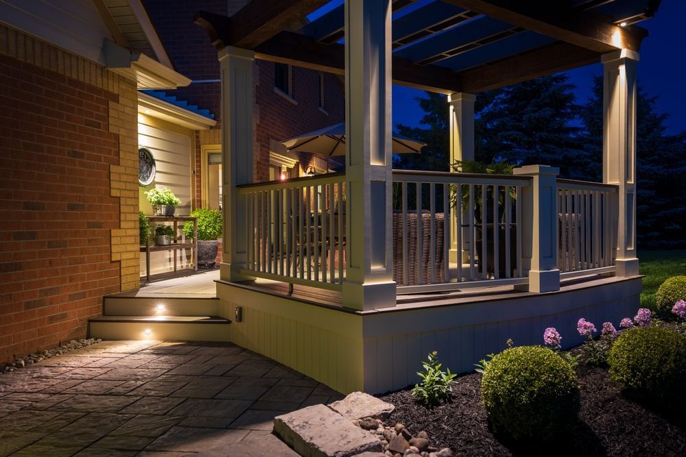 Night view of a lit outdoor patio with pergola and landscaping. Brick house on the left, patio furniture visible.