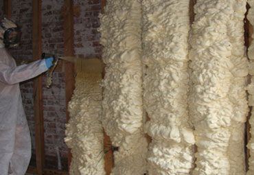 Person in protective suit spraying foam insulation onto exposed wood framing.