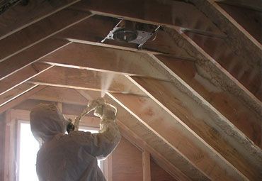 Person spraying insulation in an attic, wearing protective suit. Wooden beams visible.