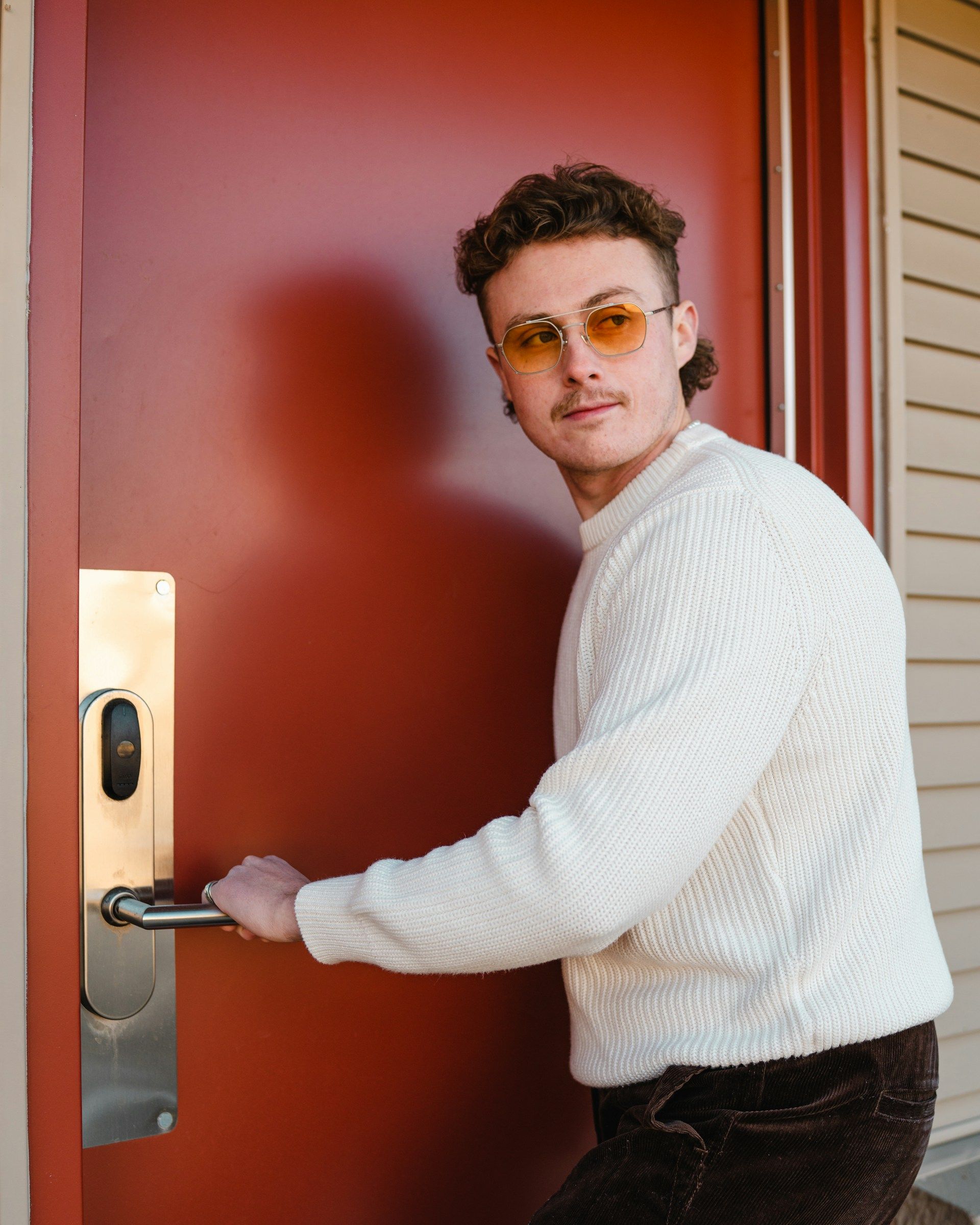 Young man wearing sunglasses unlocking a bright red apartment door, framed inside a white border