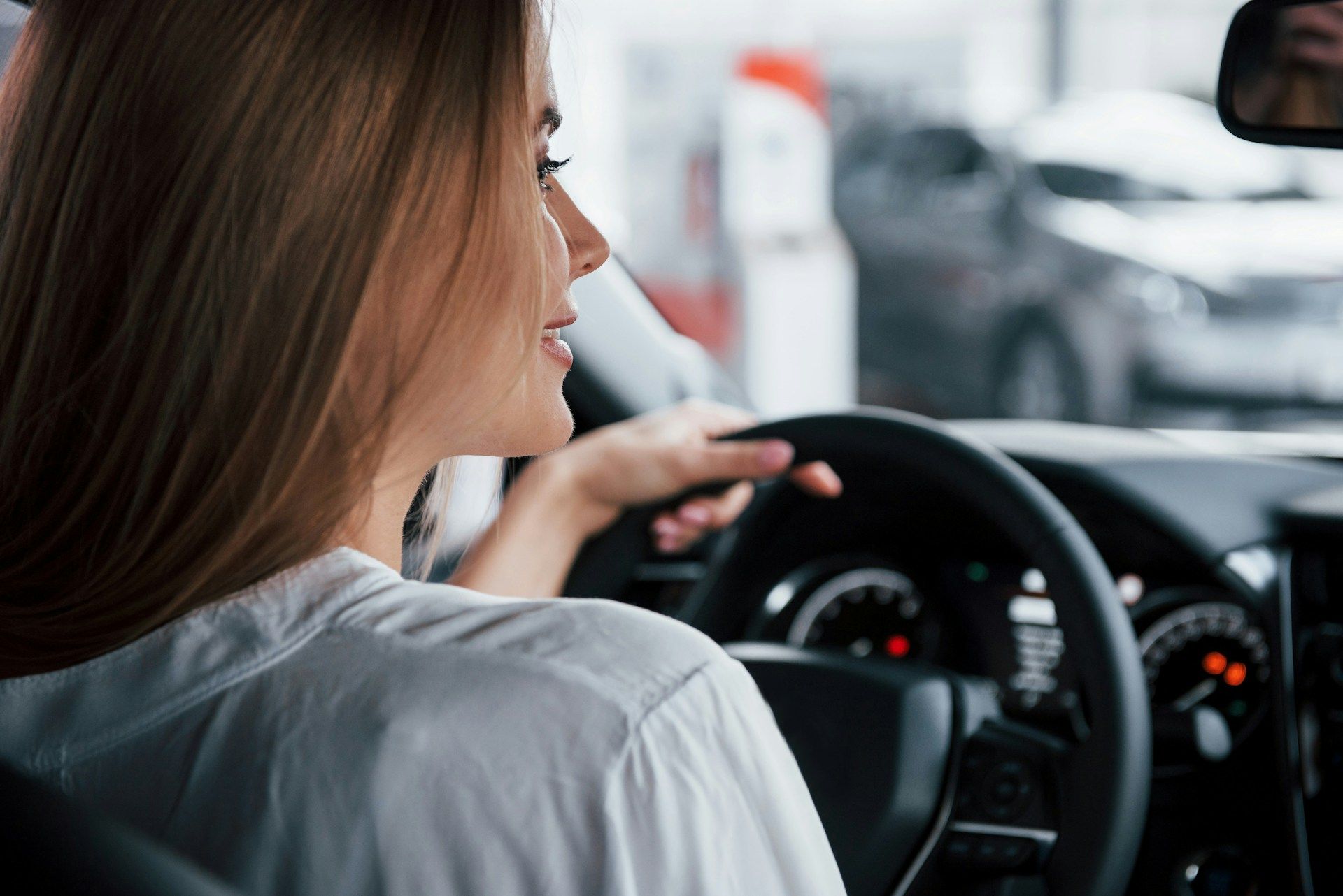 A woman with long hair seen from the side, sitting in the driver's seat of a car, looking forward with her hands on the steering wheel.