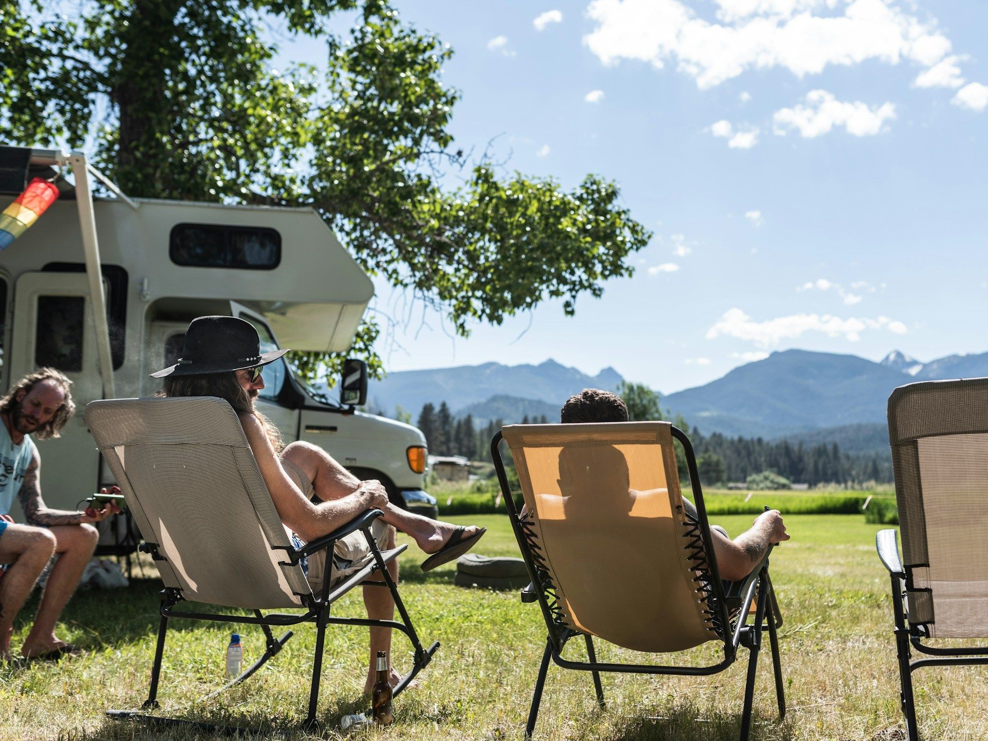 Two people relaxing in lawn chairs next to a white RV, with a campfire and scenic mountains in the background