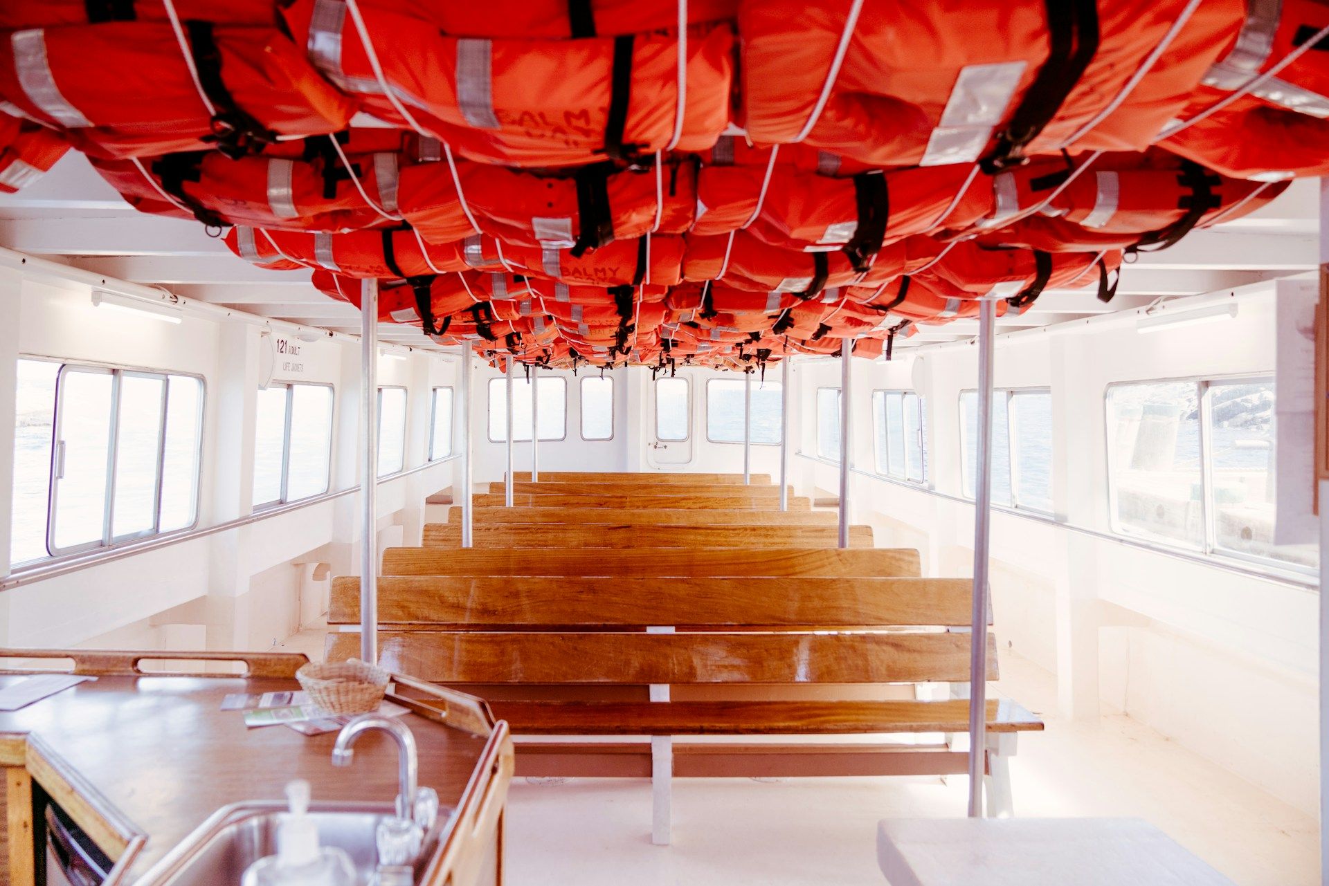 Interior view of a passenger boat showing rows of wooden benches and orange life jackets stowed in the ceiling