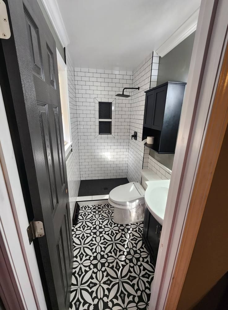 A bathroom featuring black patterned floor tiles, white subway tile shower walls, a black cabinet, and a white toilet.