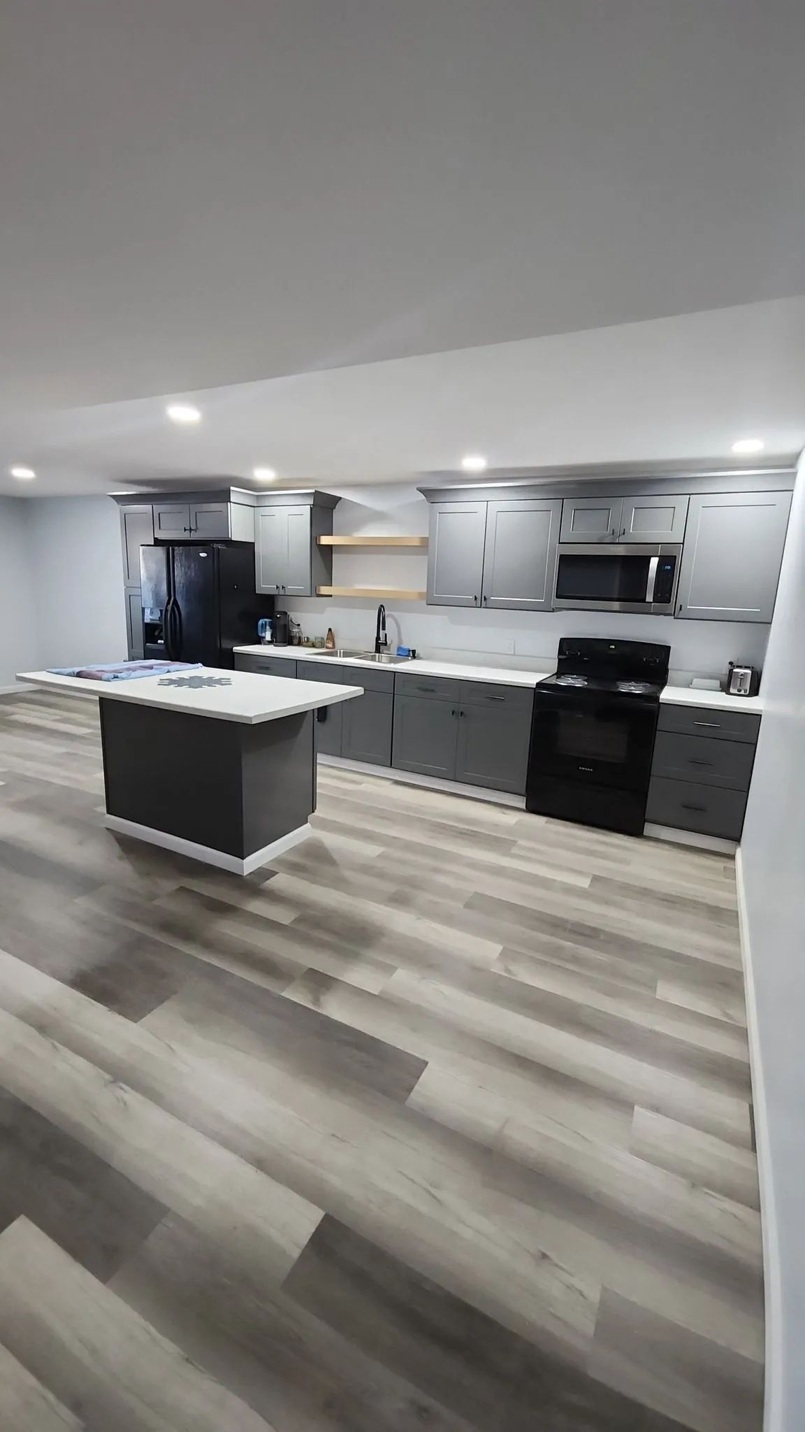 Modern kitchen with gray cabinets, white countertops, island, black appliances, and wood-look flooring.