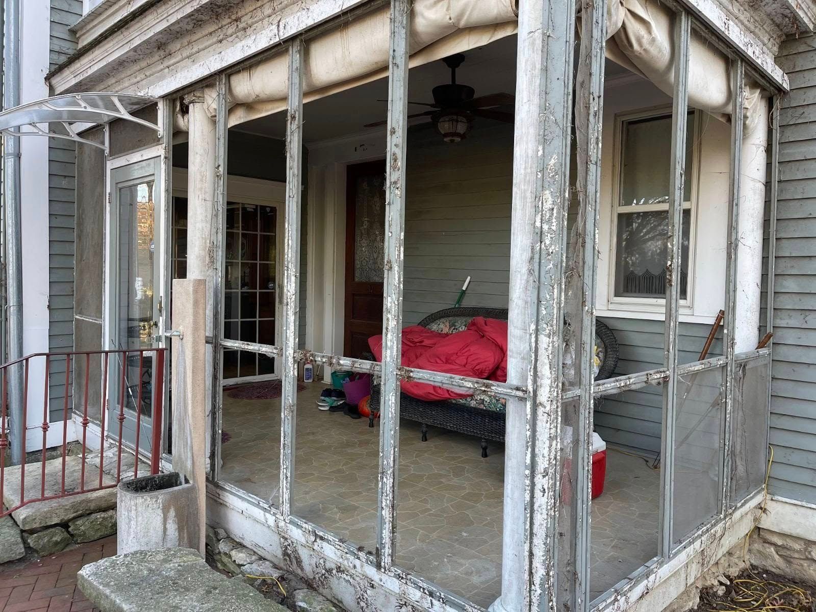 A worn, screened-in porch on the front of a house, featuring gray peeling paint, a red cushion, and a ceiling fan.