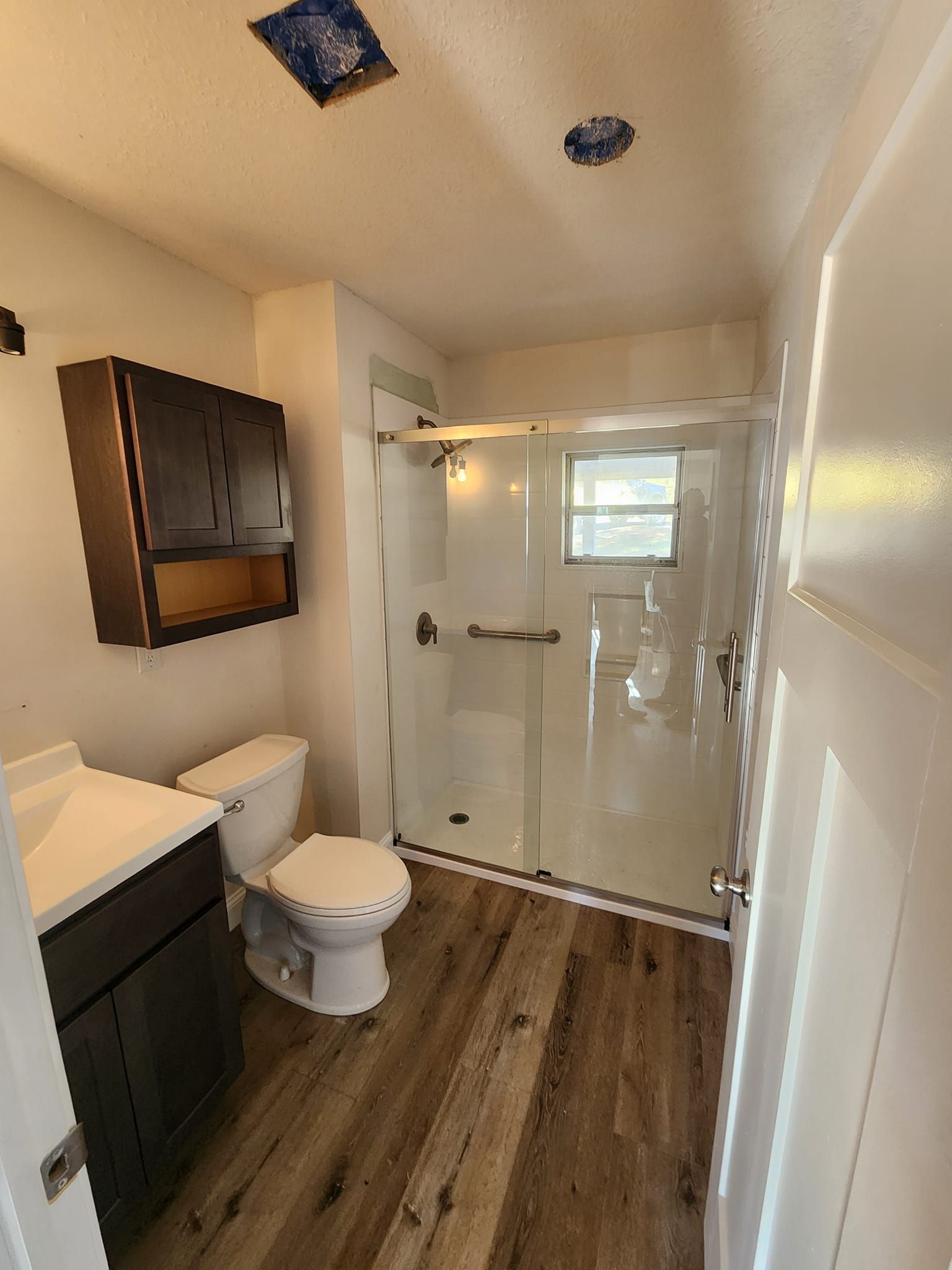 A small bathroom with a wood-look floor, a white toilet, a dark vanity, and a glass-enclosed shower.