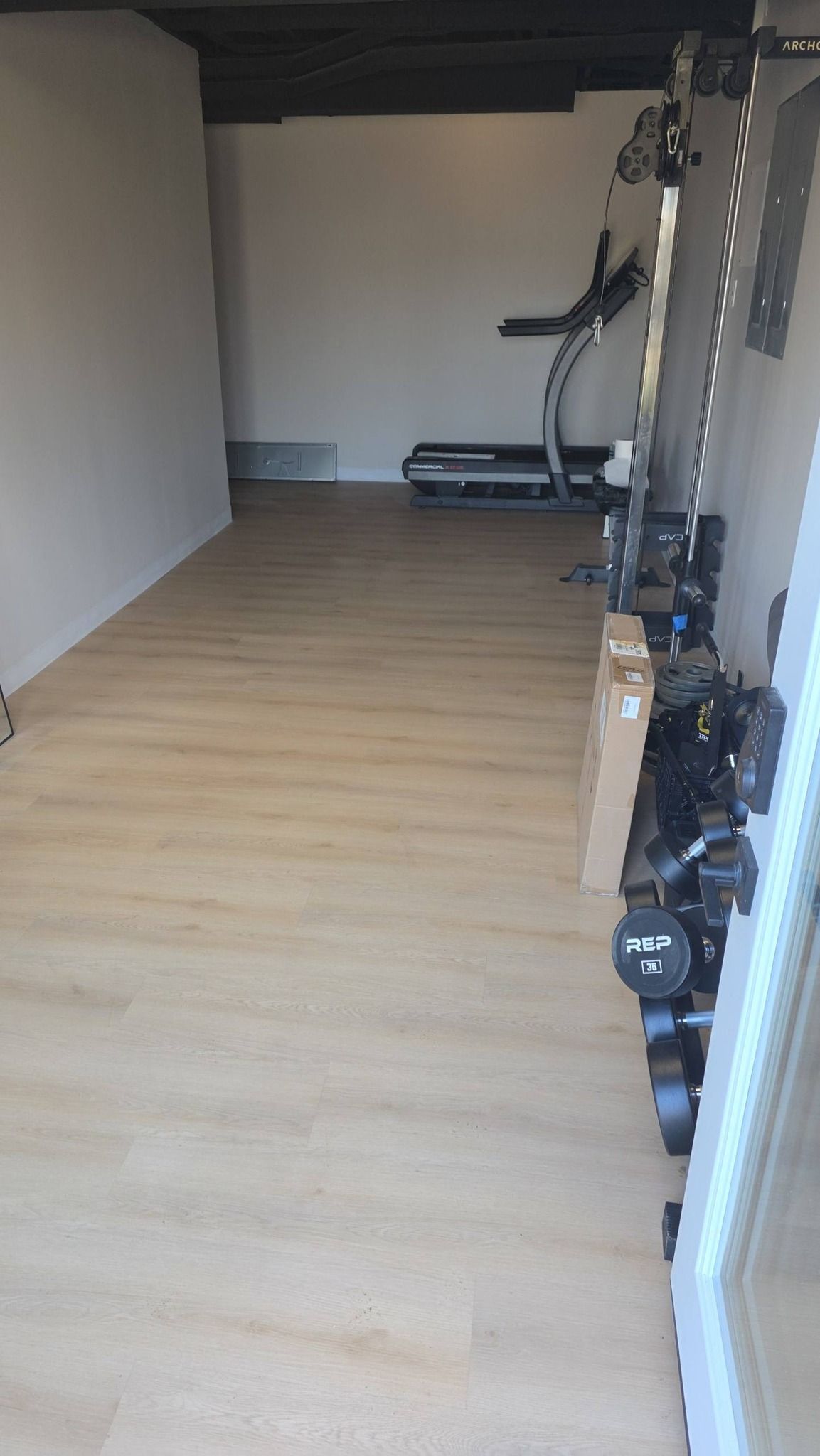 A room with light wood flooring, a treadmill in the back, and a rack of dumbbells along the right wall.
