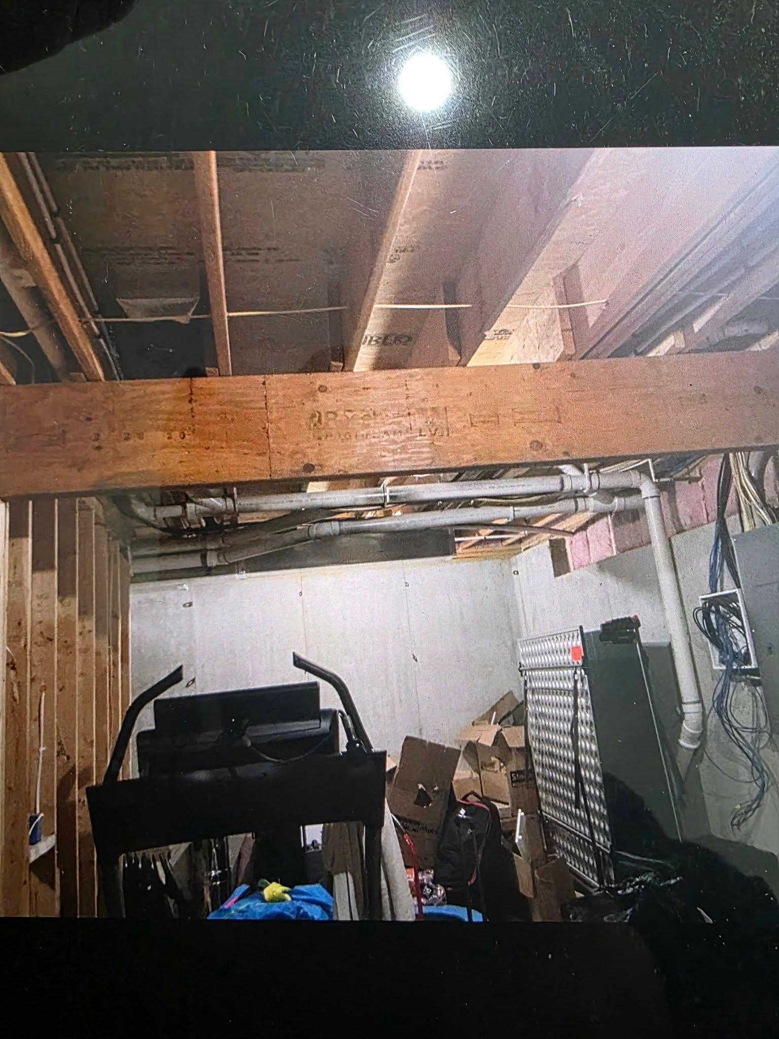 Unfinished basement with a large horizontal wooden support beam, ceiling joists, plumbing pipes, and scattered storage.