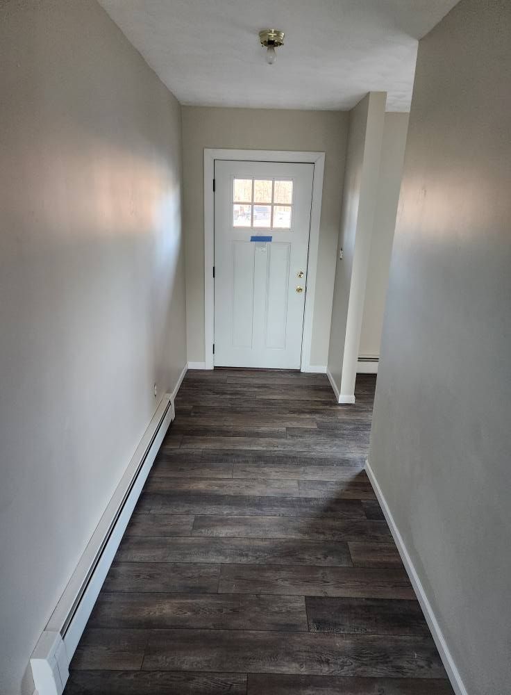 A hallway with grey walls, dark wood-look plank flooring, a white heater along the left wall, and a white front door.