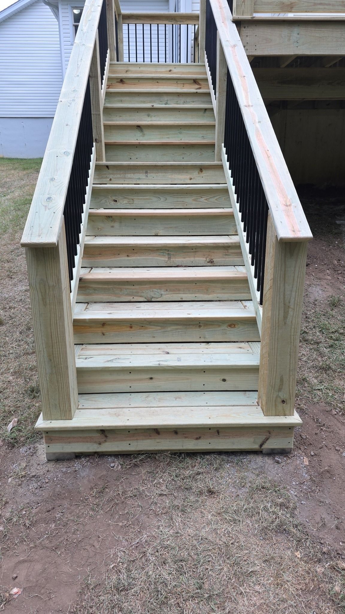 New wooden stairs with black metal railings lead up to an elevated deck next to a white house.