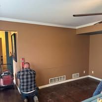 A person wearing a plaid shirt kneels on a dark floor in a room with brown walls, using a level on the wall.