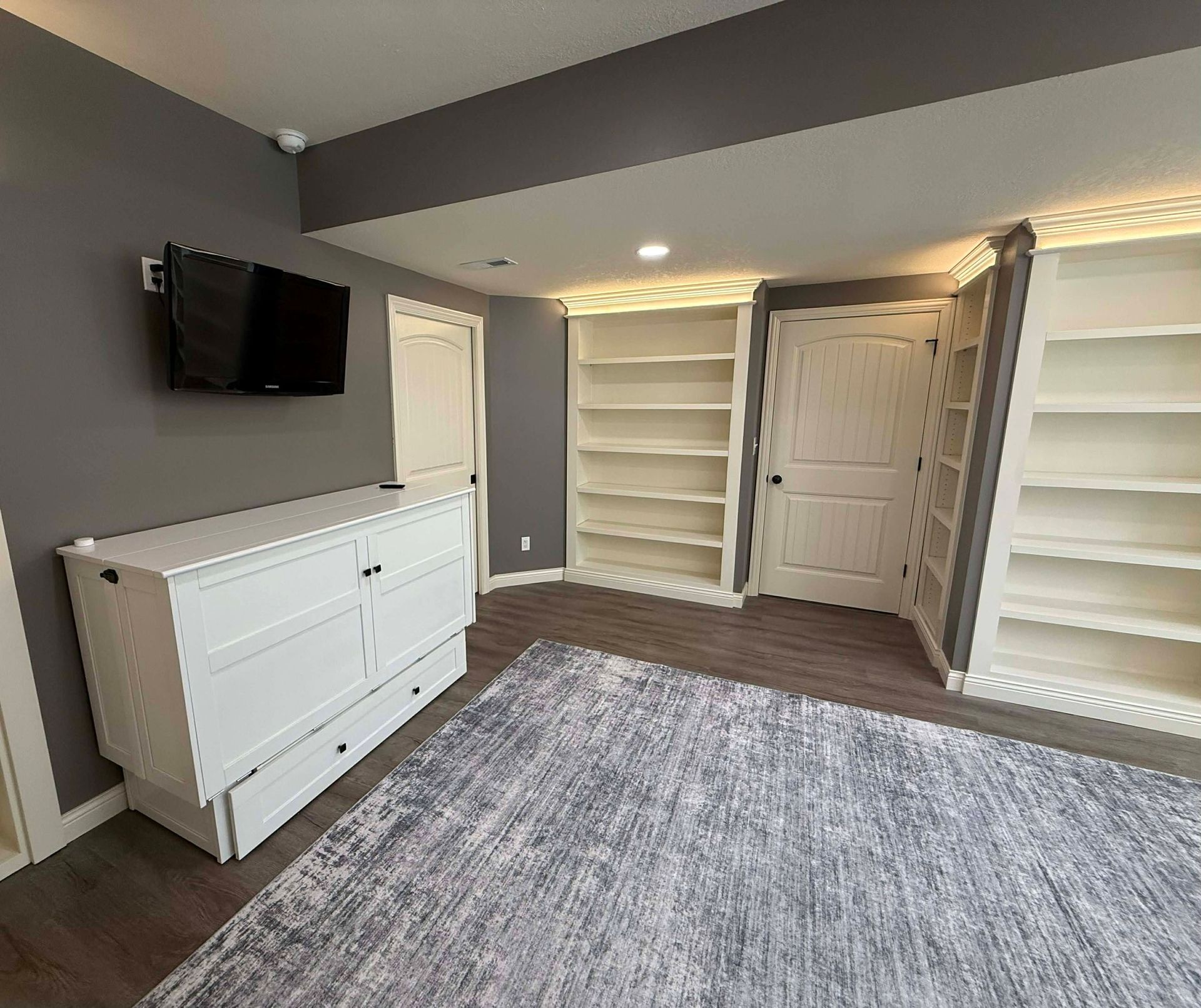 A room with dark gray walls, a white dresser topped by a TV, a rug, and white built-in bookshelves flanking a door.