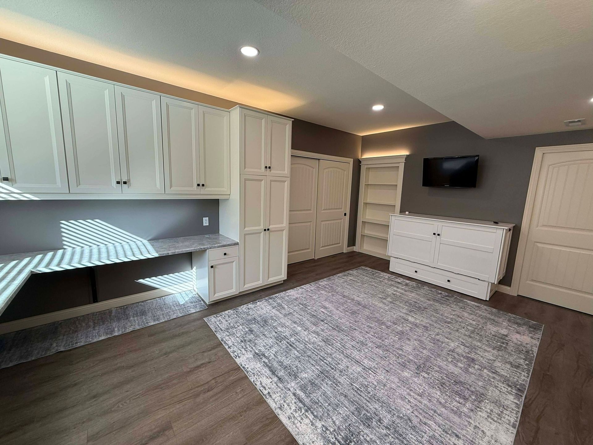 A room with white cabinetry, a large desk, a gray patterned rug, a wall-mounted TV, and a white dresser.