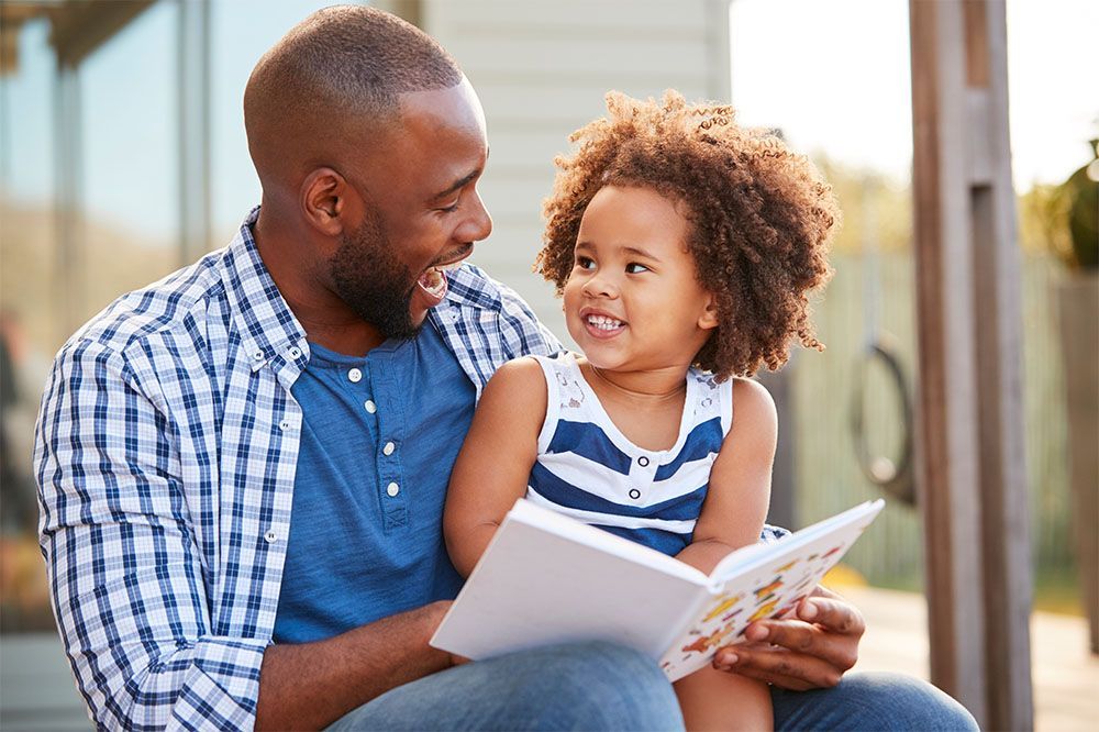 Man and young child reading a book together outdoors, smiling on a porch