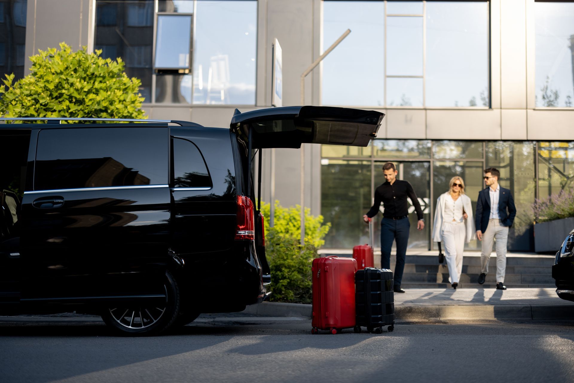 People standing beside a black van outside a modern building, with luggage in front.